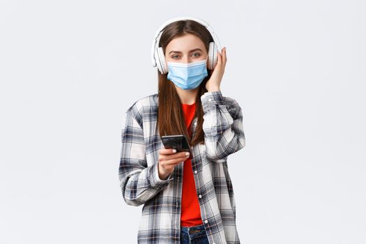 Social distancing, leisure and lifestyle on covid-19 outbreak, coronavirus concept. Cheerful smiling woman in medical mask, picking song from playlist smartphone, listen music in wireless headphones.