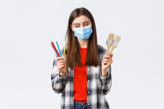 Social distancing, leisure and hobbies on covid-19 outbreak, coronavirus concept. Pleased smiling girl in medical mask start draw, look at colored pencils, show painting brushes.