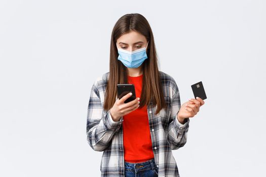 Coronavirus outbreak, working from home, online shopping and contactless payment concept. Girl in medical mask holding credit card, order clothes using mobile phone application.