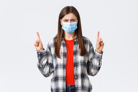 Coronavirus outbreak, leisure on quarantine, social distancing and emotions concept. Angry, outraged young woman in medical mask grimacing, looking mad as pointing up.