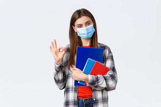 Coronavirus pandemic, covid-19 education, and back to school concept. Determined young woman, student in medical mask with notebooks and study material, show okay, no problem sign.