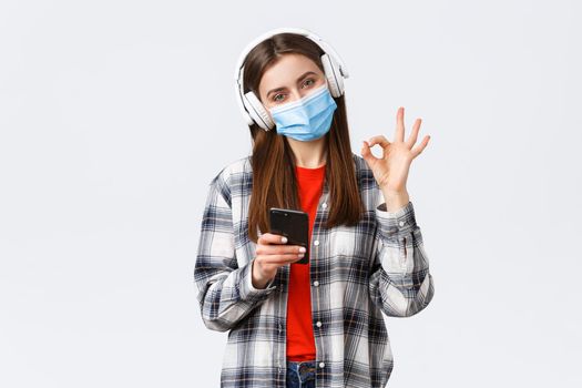 Social distancing, leisure and lifestyle on covid-19 outbreak, coronavirus concept. Satisfied attractive girl in medical mask, headphones, show okay approval as pick song from mobile phone playlist.