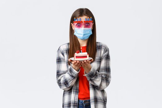 Coronavirus outbreak, lifestyle during social distancing and holidays celebration concept. Surprised or ambushed cute girl in glasses and medical mask, holding birthday cake, confused how blow candle.