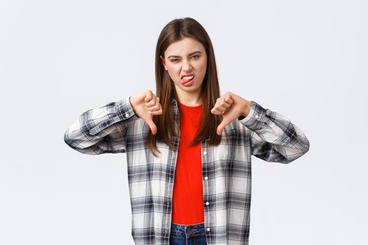 Lifestyle, different emotions, leisure activities concept. Displeased and unimpressed young picky girl in checked shirt, thumb-down and show tongue to express dislike, rate awful movie.