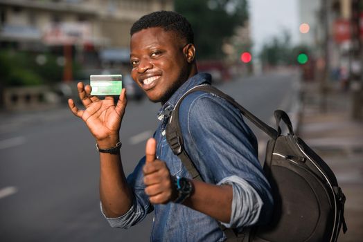 Young single traveler on the street at the platform of the station with backpack. Smiling man showing credit card, waiting for vehicle at train station for trip. Summer vacation traveling or young tourist concept