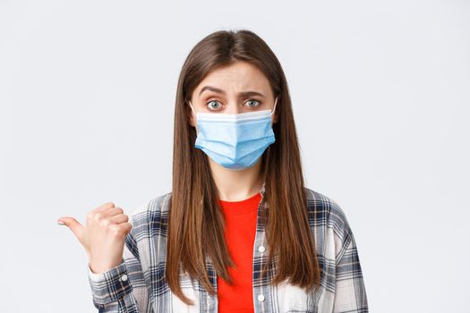Coronavirus outbreak, leisure on quarantine, social distancing and emotions concept. Skeptical, unimpressed pretty woman in medical mask pointing thumb left and frowning doubtful.
