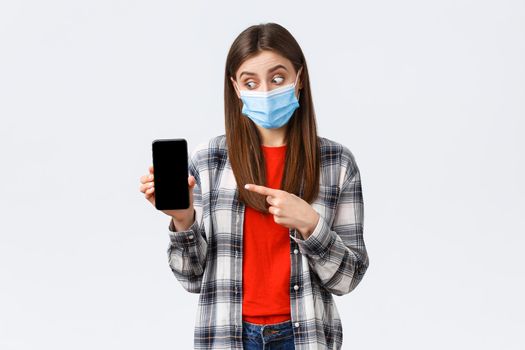 Different emotions, covid-19, social distancing and technology concept. Excited and enthusiastic good-looking girl in medical mask, pointing looking at mobile phone display, promote app.