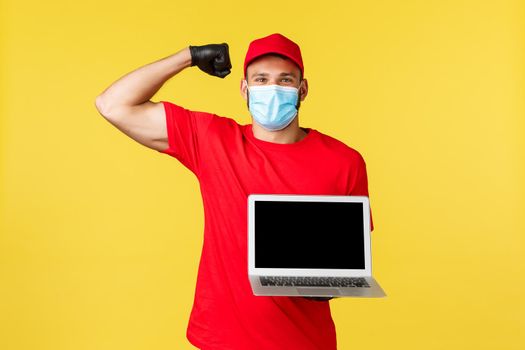 covid-19, safe shopping, express delivery and order tracking concept. Strong handsome courier in red uniform and medical mask, flex biceps and showing laptop screen, smiling.