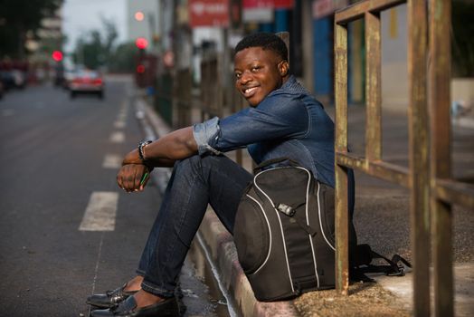 young happy traveler man with backpack, sitting alone on the street in the evening, waiting for a car or bus at the bus stop,