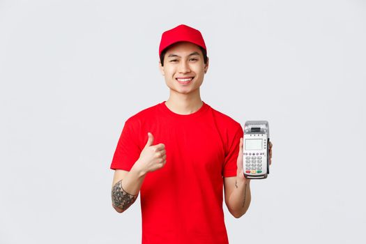 Cheerful smiling asian delivery guy recommend using credit cards, no cash payment during covid 19 self-quarantine, showing payment terminal POS and thumb-up, approve contactless purchase.