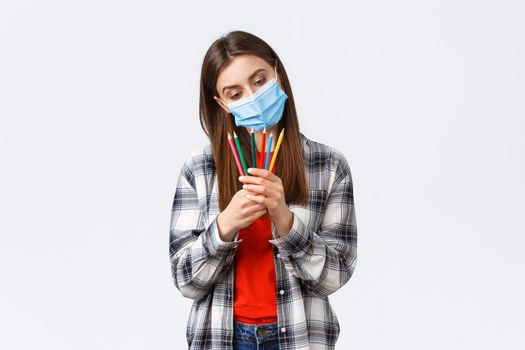 Social distancing, leisure and hobbies on covid-19 outbreak, coronavirus concept. Thoughtful cute, dreamy woman in medical mask decide which colored pencil choose, pondering what draw.