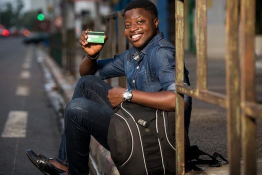 Young single traveler on the street at the platform of the station with backpack. Smiling man showing credit card, waiting for vehicle at train station for trip. Summer vacation traveling or young tourist concept