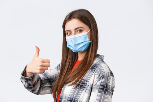 Coronavirus outbreak, leisure on quarantine, social distancing and emotions concept. Cheerful young woman in medical mask stand in profile, turn to camera and thumb-up in approval.
