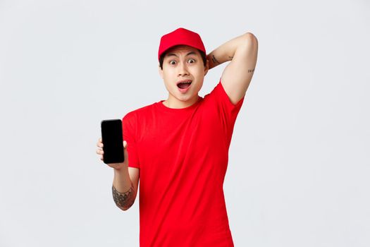 Nervous and troubled asian delivery guy in red cap and t-shirt uniform, gasping anxious, look confused and puzzled showing smartphone screen bad news, mixed-up order, stand grey background.
