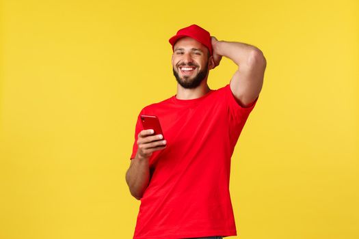 Contactless payment, online delivery, internet shopping and parcel tracking concept. Carefree young bearded guy, courier in red cap and t-shirt, laughing as using mobile phone, yellow background.