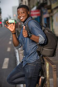 Young single traveler on the street at the platform of the station with backpack. Smiling man showing credit card, waiting for vehicle at train station for trip. Summer vacation traveling or young tourist concept