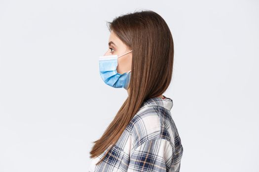 Coronavirus outbreak, leisure on quarantine, social distancing and emotions concept. Profile of serious-looking young pretty woman in medical mask standing in line, white background.