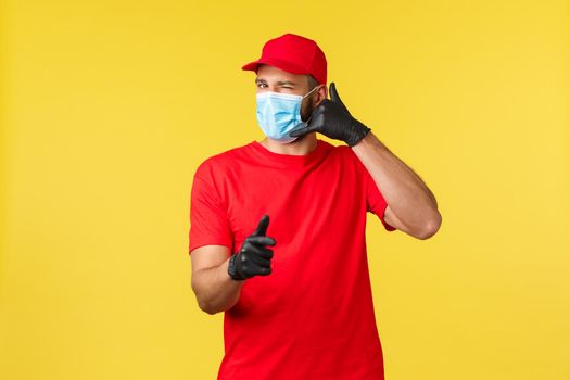 Express delivery during pandemic, covid-19, safe shipping, online shopping concept. Friendly courier in red uniform, medical mask and gloves, wink and ask call service, pointing camera.