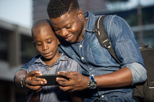 Father and son sitting in the street waiting for car, and looking on cell phone.