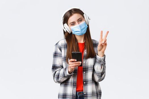 Social distancing, leisure and lifestyle on covid-19 outbreak, coronavirus concept. Cute silly teenage girl in medical mask and headphones, show peace sign, relaxing with music, hold mobile phone.