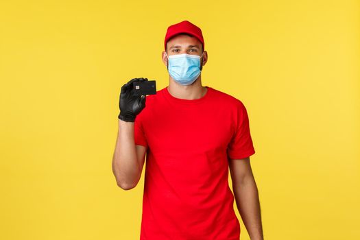 Express delivery during pandemic, covid-19, safe shipping, online shopping concept. Young guy courier in medical mask and gloves, explain use of contactless paying with credit card.