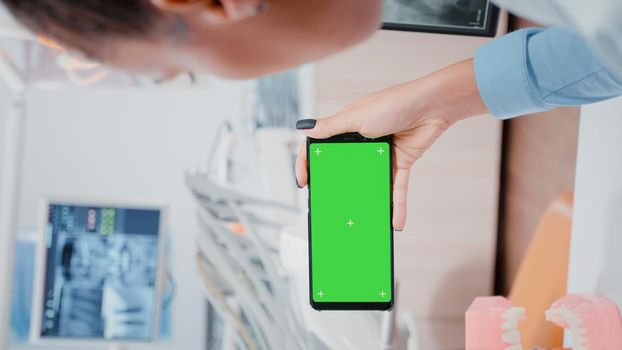 Vertical video: Woman holding smartphone with horizontal green screen at oral care clinic. Dentist looking at chroma key with isolated background and mockup template for dental checkup