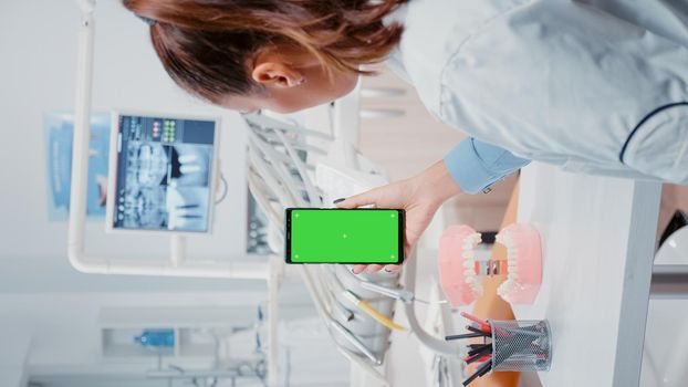Vertical video: Woman vertically holding mobile phone with green screen in dental cabinet. Stomatologist looking at chroma key and isolated mockup template on smartphone for oral care at office