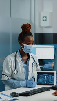 Doctor holding tablet with human body analysis for checkup visit with patient, wearing face masks against covid 19 epidemic. Medic explaining skeleton illustration for osteopathy care.