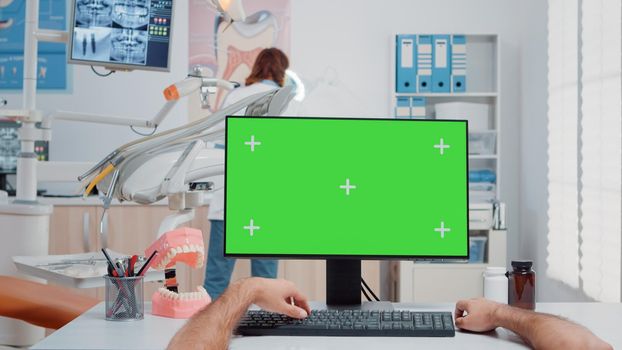 POV of assistant working with green screen on computer at dentist office. Man using monitor with chroma key and isolated mockup template for dentistry and teethcare in dental cabinet