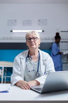 Portrait of general practitioner with stethoscope and white having laptop on desk in medical office. Woman doctor sitting in cabinet with computer and teechnology for healthcare service.