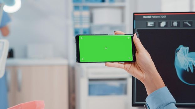 Close up of orthodontist holding horizontal green screen on smartphone in oral care office. Dentist looking at mockup template and isolated background for teethcare and dentistry.