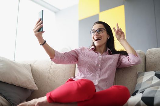Low angle of pretty young female show peace sign and take selfie on smartphone. Talk to friend or family on video call. Technology, fun, leisure concept