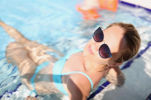 Top view of happy young female sit in pool wearing trendy glasses. Relaxing atmosphere on vacation, sunny day. Holiday, resort, hotel, time alone concept