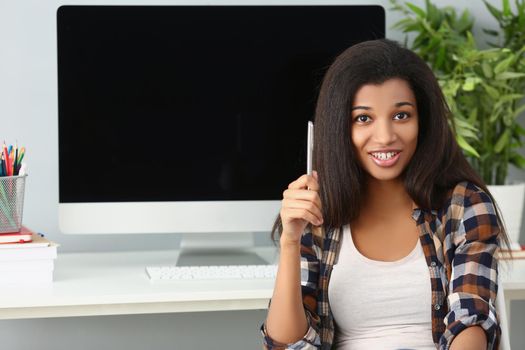 Portrait of afro american young lady sit in office with computer device behind on desk. Cute woman with curly hairstyle hold silver pen. Secretary concept