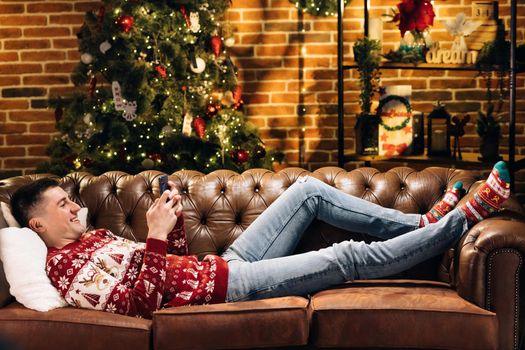 Attractive guy looking attentively favorite film. Good-looking Caucasian guy lying and watching TV using mobile phone. Happy student jumping on couch in decorated apartment in evening after work.