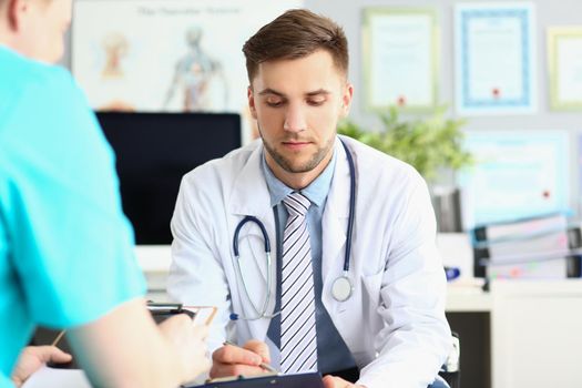 Portrait of young intern man in uniform and stethoscope, work meeting in doctor office. Listen to instruction of professional, make notes. Hospital concept