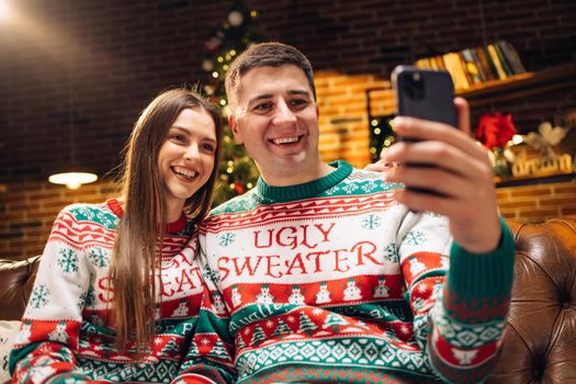 Couple in winter sweaters record video of New Year's greetings for friends and family at home. Family using smart phone talking to family congratulates Merry Christmas by video call conference app