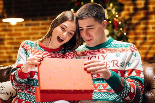 Portrait of a romantic couple opening a present gift box in the evening near decorated xmas tree. Happy man is making christmas gift to his beloved woman. Concept of holidays, romance, surprise.