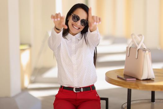 Portrait of cheerful happy young woman in stylish outfit show some symbols with hands. Attractive lady sit outdoors, gesture on camera. Trendy, joy concept