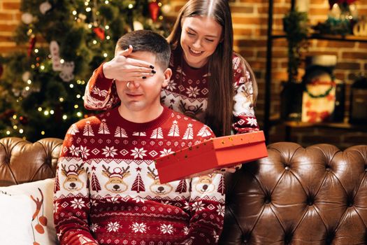 Smiling young 30s woman covering eyes of smiling curious husband, giving wrapped box with Christmas gift near decorated festive tree, New Year winter holidays family celebration. Holiday miracle.