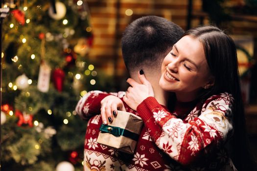 Happy woman embracing her boyfriend and hugging him near decorated glowing fir tree. Romantic winter story. Happy wife feels grateful hugs husband express gratitude for New Year gift.