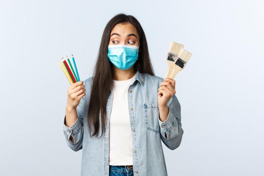 Social distancing lifestyle, covid-19 pandemic, self-isolation hobbies and leisure concept. Excited creative asian girl in medical mask showing coloured pencils and painting brushes, want draw.