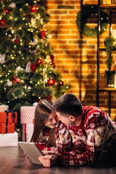 Cheerful caucasian couple man and woman resting at home near glowing new year tree and typing on tablet looking for xmas presents online on holiday sales. Buy online, sale, modern tech overuse concept