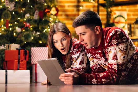 Caucasian couple lying home near Christmas tree background. Beautiful woman with long hair holds digital tablet and emotionally tells funny stories.