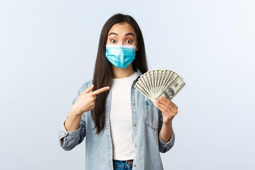Social distancing lifestyle, covid-19 pandemic business and employement concept. Excited female asian employee suggest you earn lots money, pointing at cash dollars, wear medical mask.