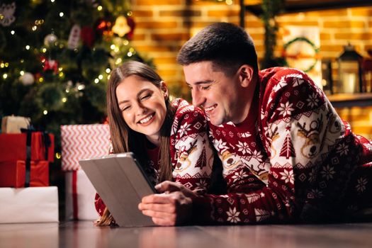 Happy caucasian couple lying home near Christmas tree background. Beautiful woman with long hair holds digital tablet and emotionally tells funny stories.