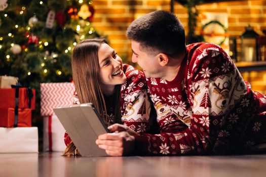 Caucasian family couple choosing Christmas present online on tablet while lying at cozy home. Husband and wife looking for xmas gift on tablet computer device. New Year spirit. Holidays celebration.