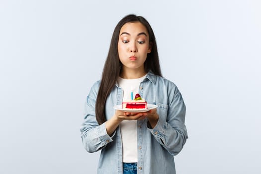 Social distancing lifestyle, covid-19 pandemic, celebrating holidays during coronavirus concept. Playful cute and silly asian girl pouting, blowing candle on birthday cake, making wish.