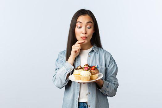 Social distancing lifestyle, covid-19 pandemic, celebrating holidays during coronavirus concept. Delighted and temting cute asian girl staring with desire at plate of cupcakes.