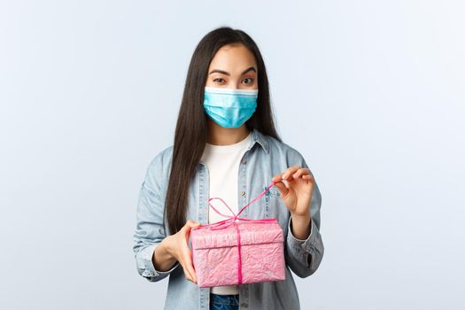 Social distancing lifestyle, covid-19 pandemic, celebrating holidays during coronavirus concept. Intrigued happy asian woman in medical mask unwrap gift box wonder what inside.